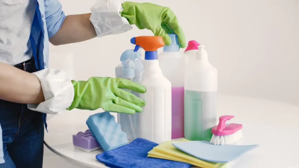Pessoa com luvas verdes organizando produtos de limpeza sobre uma mesa branca, preparando-se para mostrar como limpar rejuntes com eficiência.