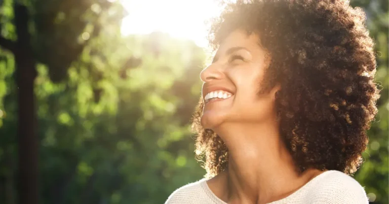 Mulher negra sorrindo ao ar livre, olhando para o alto com o sol ao fundo.