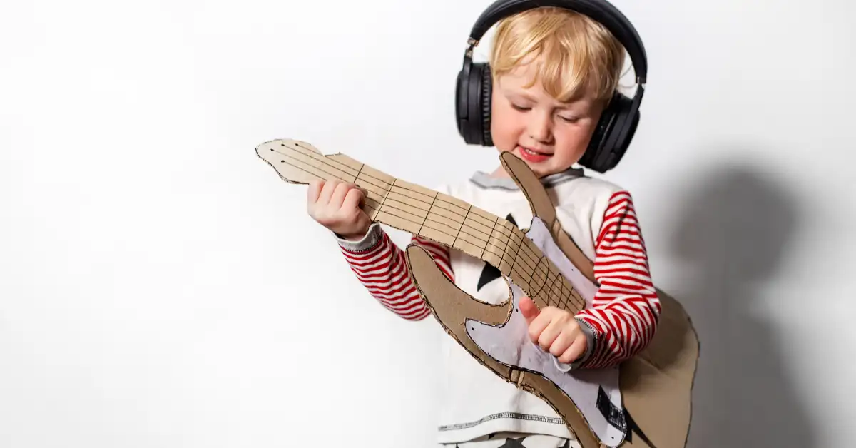 Criança usando fones de ouvido e tocando uma guitarra de papelão, em uma atividade educativa em casa que estimula a criatividade.
