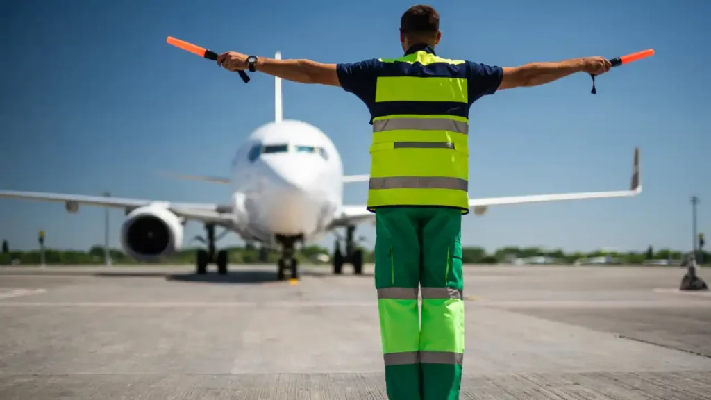 Membro da equipe de solo orienta aeronave no pátio sob céu azul, representando operações seguras e práticas de aviação sustentável.