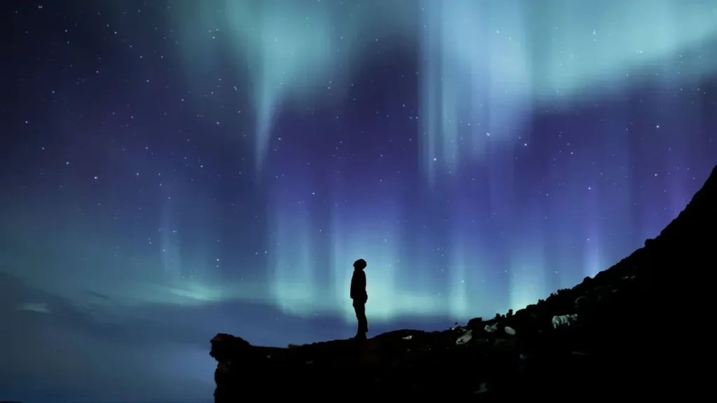 Destinos congelados que brilham no escuro – Silhueta de pessoa sobre penhasco rochoso observando a aurora boreal em noite estrelada