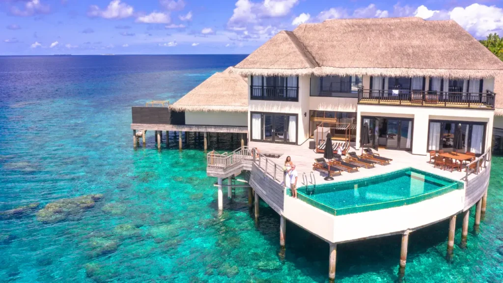 Vista aérea de uma villa luxuosa sobre palafitas em ilhas privadas com piscina de borda infinita, cercada por águas cristalinas e azuis do oceano.