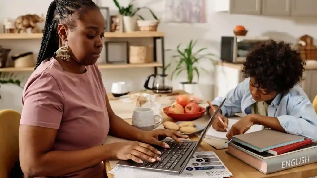 Mãe e filho realizando atividades educativas em casa, com uso de laptop, livros e cadernos em uma mesa de estudos.