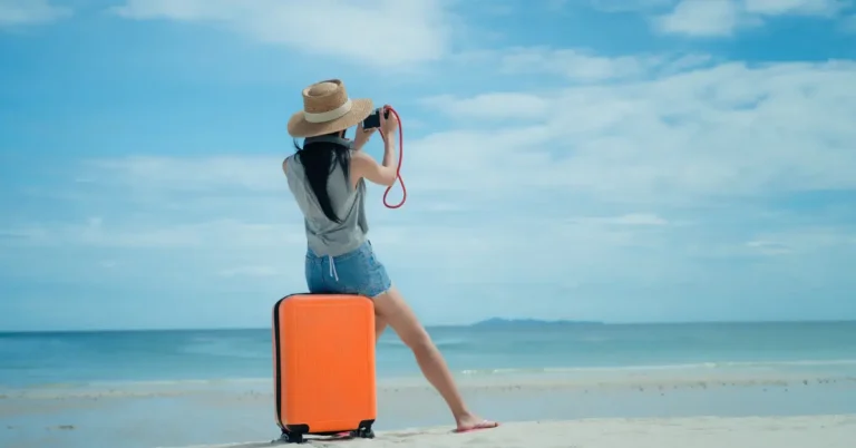 Pessoa em pé na praia com uma mala laranja, tirando foto do mar — inspiração para "o que levar na mala para passar 7 dias no litoral"