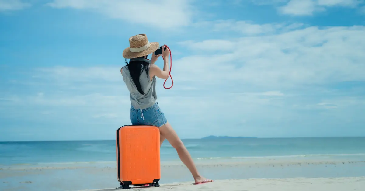 Pessoa em pé na praia com uma mala laranja, tirando foto do mar — inspiração para "o que levar na mala para passar 7 dias no litoral"