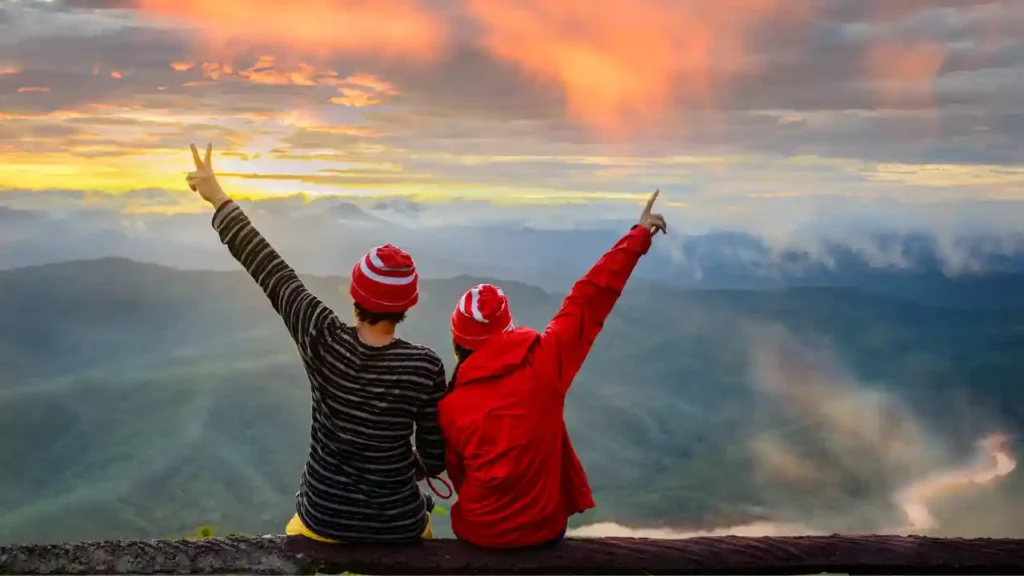 Dois viajantes com gorros listrados apreciando o pôr do sol nas montanhas — inspiração para o que levar na mala para viagem de inverno.