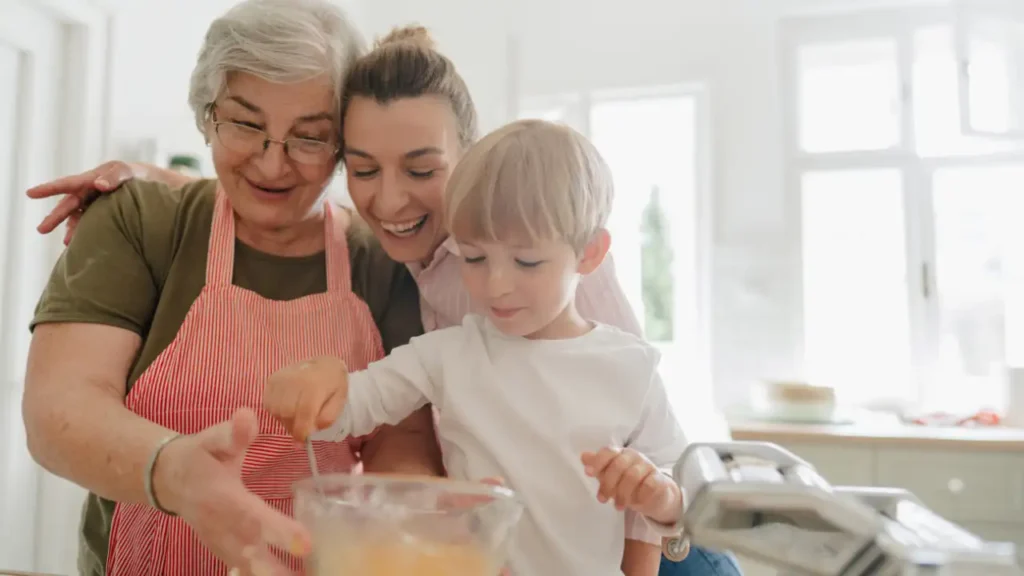Família reunida na cozinha preparando receitas simples da vovó, com criança mexendo a mistura em uma tigela enquanto os adultos acompanham com afeto.