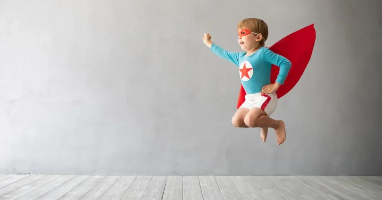 Criança pulando no ar vestida com fantasia de super-herói, com capa vermelha e camiseta azul com estrela branca em um círculo vermelho — ideal para um aniversário temático.