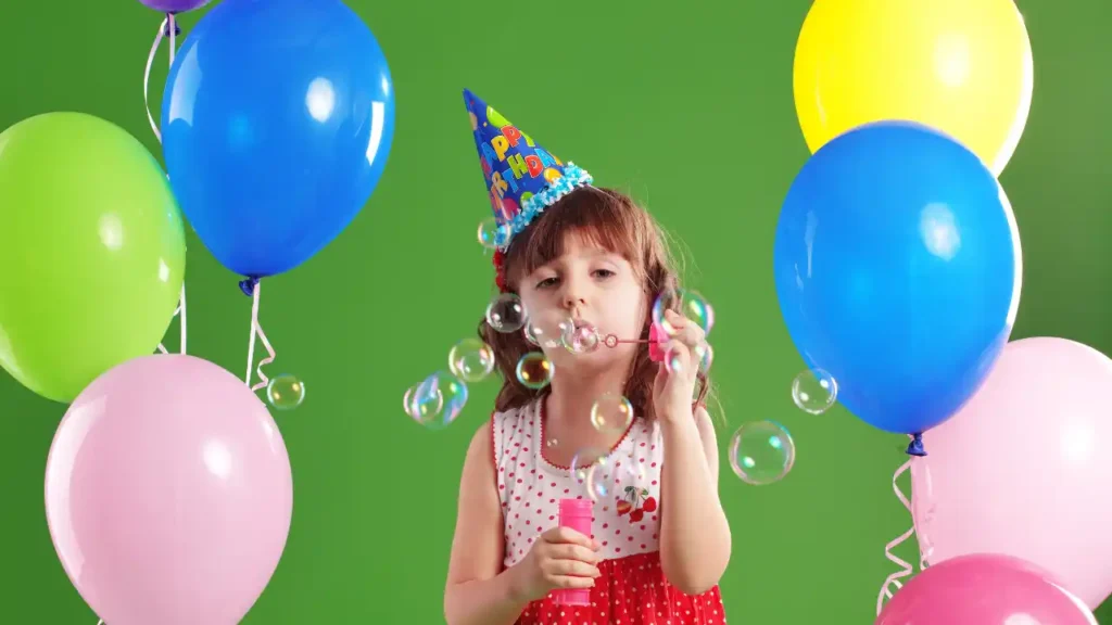 Criança soprando bolhas de sabão em meio a balões coloridos, usando chapéu de festa com a frase "Happy Birthday" — celebração alegre de um aniversário temático.