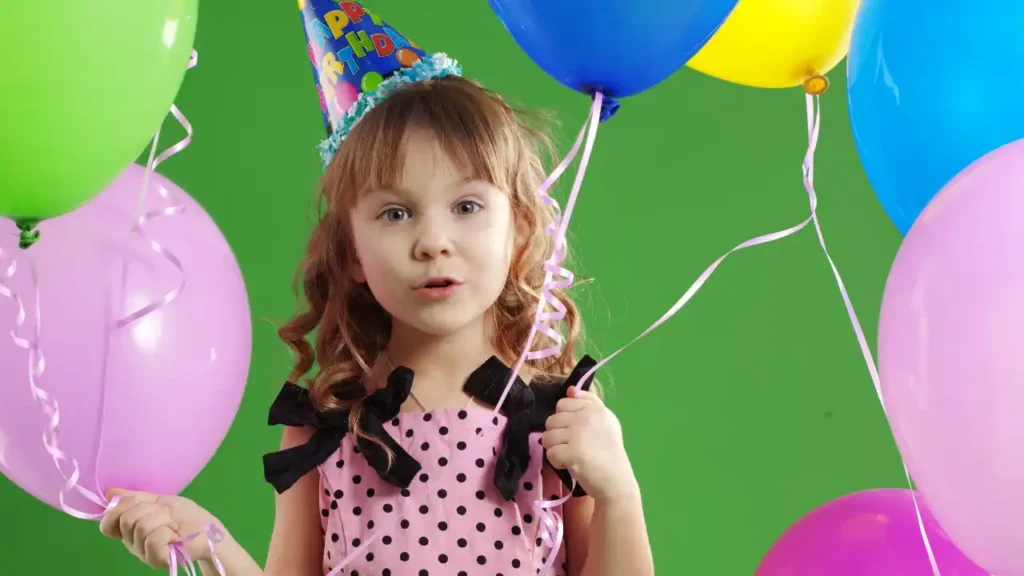 Criança com vestido rosa de bolinhas pretas segurando balões coloridos e usando chapéu de festa que diz "Happy Birthday" — um momento encantador de um aniversário temático.