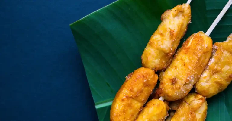 Espetinhos de banana frita crocante sobre folha de bananeira, com aparência dourada e textura crocante, servidos em fundo azul escuro.
