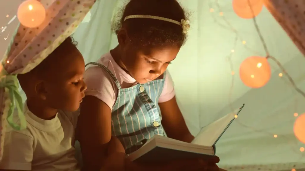 Duas crianças explorando um cantinho de leitura infantil dentro de uma cabana decorada com luzes, em clima acolhedor e mágico.