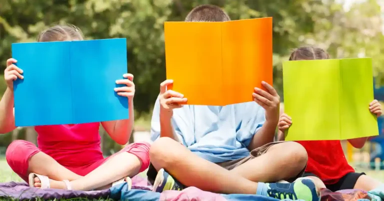 Três crianças envolvidas em leitura individual ao ar livre, formando um cantinho de leitura infantil sobre uma manta cercada pela natureza.