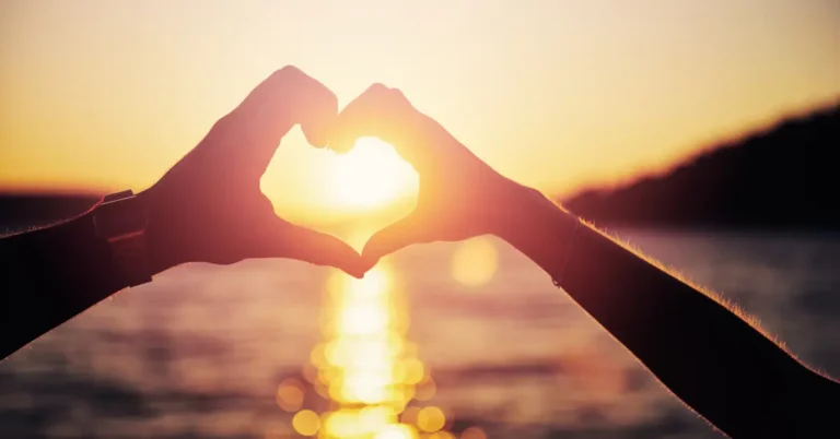Mãos formando um coração contra o pôr do sol, simbolizando afeto e conexão, cenário ideal para rituais com ervas que atraem amor.