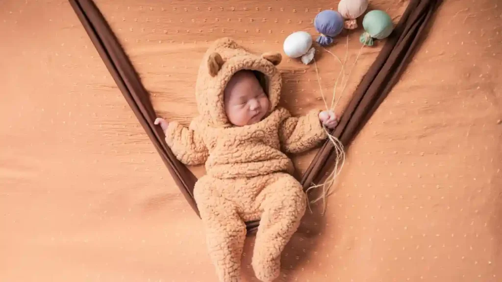 Bebê vestido com fantasia de urso marrom, deitado em uma rede de tecido, segurando balões coloridos — uma das fotos criativas de bebês que mistura fantasia e cenário lúdico.