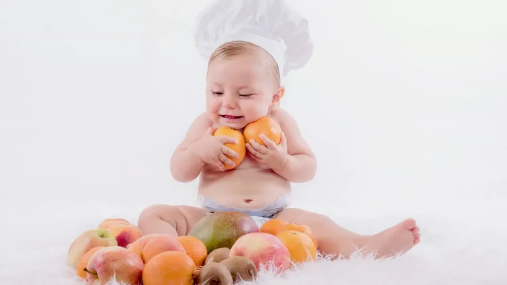 Bebê com chapéu de chef sentado sobre superfície branca felpuda, cercado por frutas variadas — uma das fotos criativas de bebês com tema culinário.