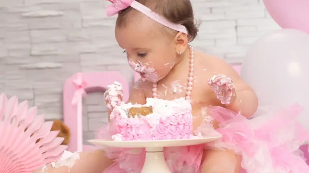 Bebê com tutu rosa e tiara sentada diante de bolo decorado, rodeada por balões e enfeites cor-de-rosa — uma das fotos criativas de bebês em clima de celebração.