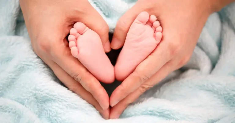 Mãos adultas seguram com delicadeza os pés de um recém-nascido envolto em manta azul, formando um coração — uma das fotos de recém-nascido que simbolizam amor e acolhimento.