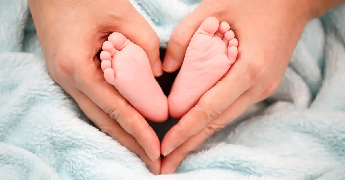 Mãos adultas seguram com delicadeza os pés de um recém-nascido envolto em manta azul, formando um coração — uma das fotos de recém-nascido que simbolizam amor e acolhimento.