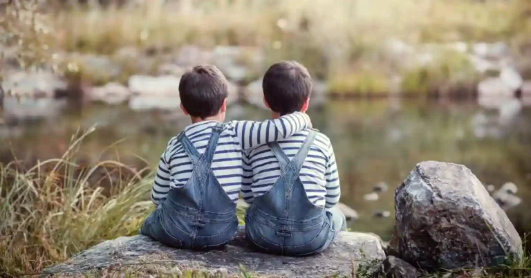 Dois irmãos em roupas combinando sentados em uma pedra à beira do lago, simbolizando conexão e carinho — inspiração para escolher nomes para gêmeos.