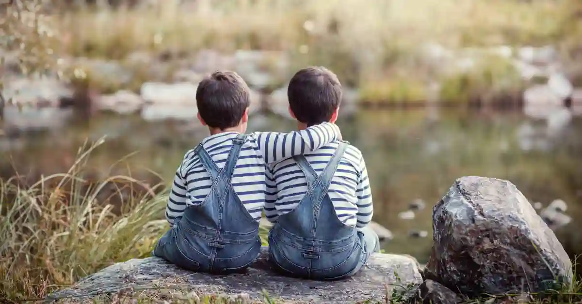 Dois irmãos em roupas combinando sentados em uma pedra à beira do lago, simbolizando conexão e carinho — inspiração para escolher nomes para gêmeos.