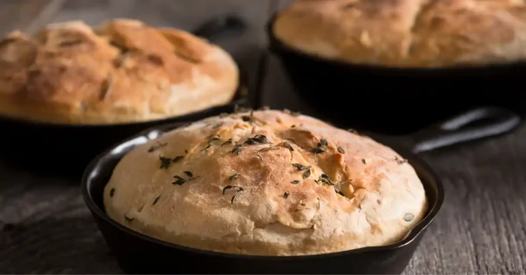 Três pães de frigideira com aveia recém-assados em frigideiras de ferro, com crosta dourada e cobertura de ervas, sobre uma bancada rústica.