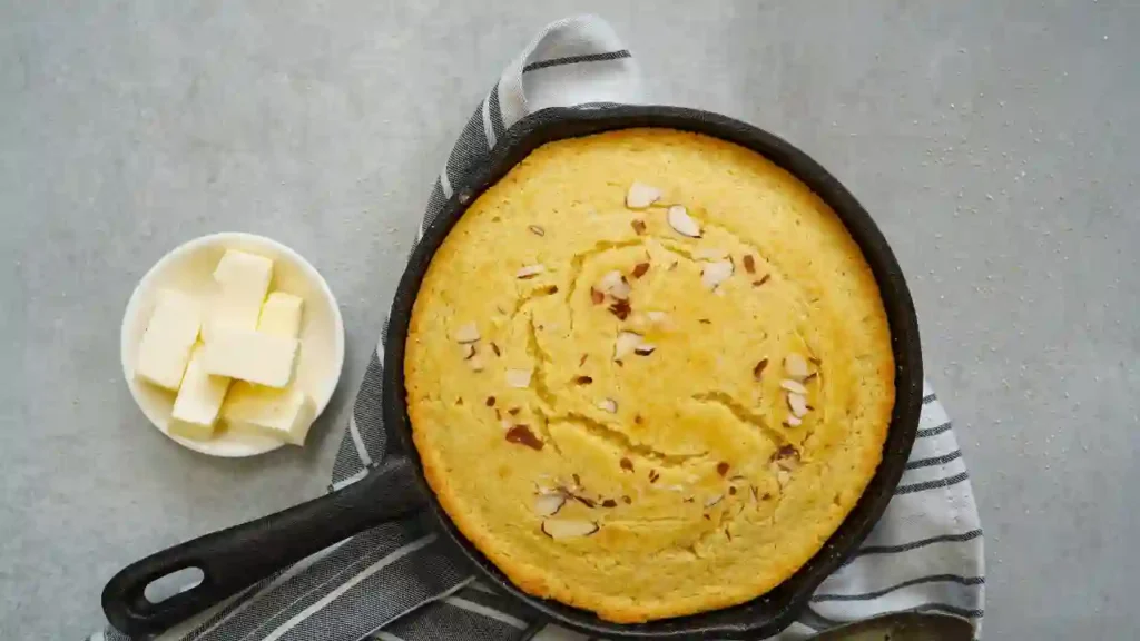 Pão de frigideira com aveia assado em frigideira de ferro, decorado com lâminas de amêndoa, ao lado de um pote branco com pedaços de manteiga sobre pano listrado.