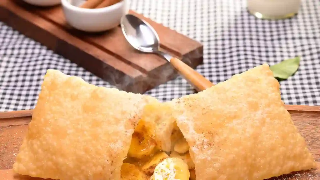 Pastel de banana frito aberto, revelando recheio cremoso, com açúcar de confeiteiro polvilhado e elementos culinários ao fundo.