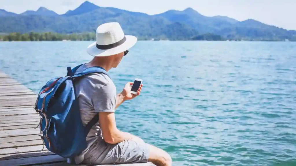 Pessoa sentada em um píer de madeira contemplando um lago calmo com montanhas ao fundo, usando chapéu branco e mochila azul, possivelmente refletindo sobre planejamento de viagens.