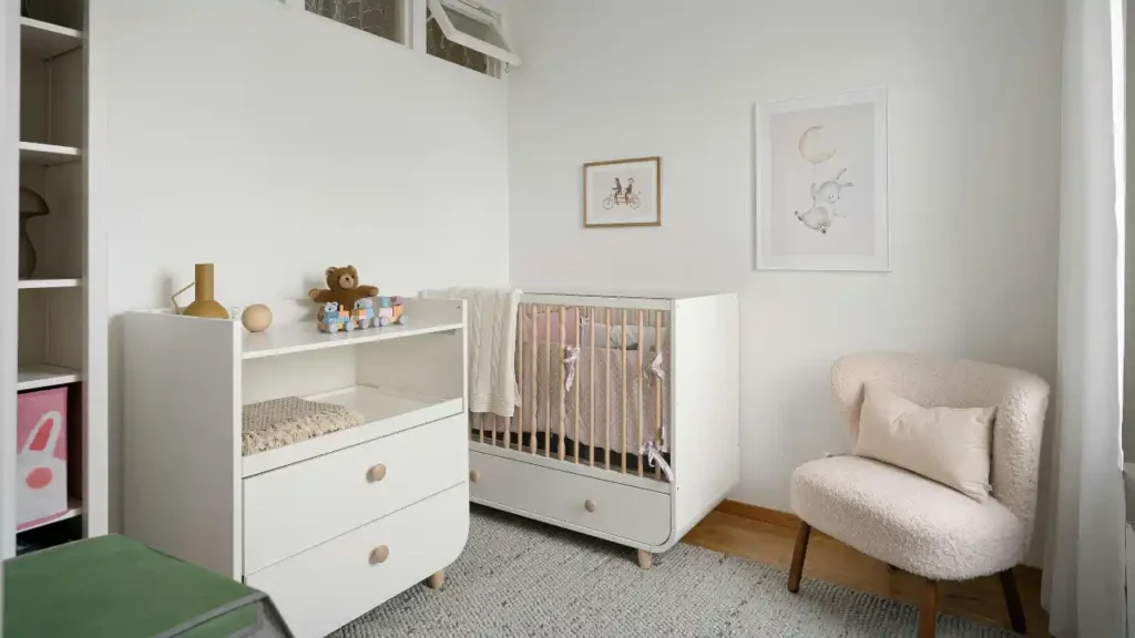 Quarto de bebê com berço branco de madeira, cômoda com brinquedos, poltrona estofada bege e quadros decorativos infantis na parede.