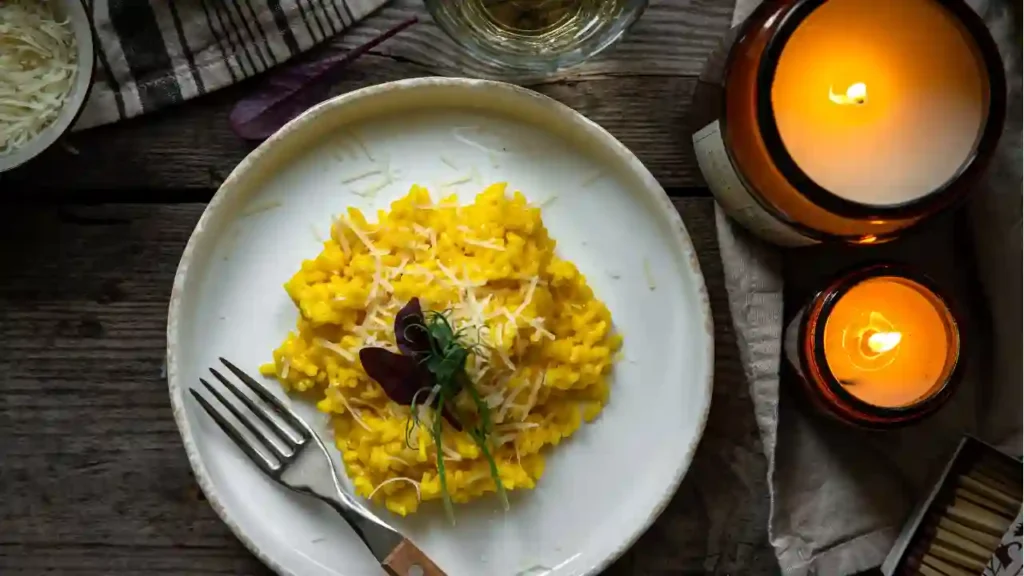 Prato de risoto quatro queijos com açafrão, decorado com ervas frescas e queijo ralado, acompanhado de taça de vinho branco, velas acesas e ambiente acolhedor sobre superfície de madeira.