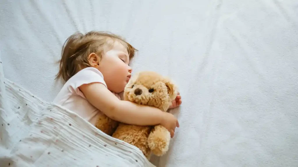 Criança dorme tranquilamente na cama, abraçada a um ursinho de pelúcia, coberta por um edredom branco com estampa de estrelas — retratando o sono do bebê.