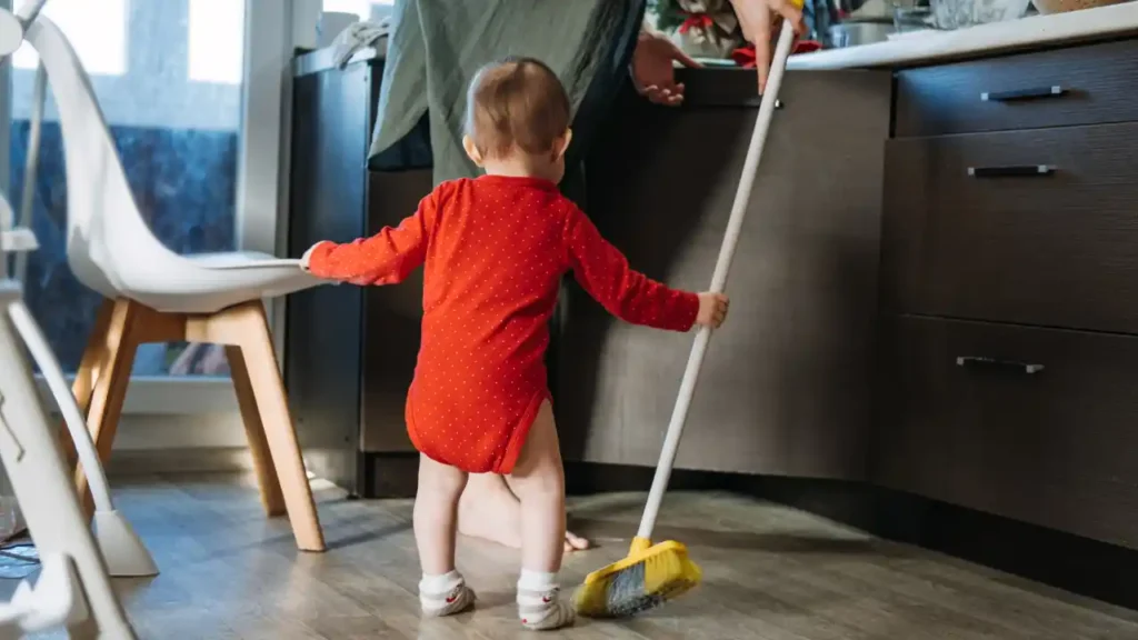 Criança pequena em roupa vermelha com bolinhas brancas segura uma vassoura na cozinha — uma cena que ilustra a verdade por trás da culpa.