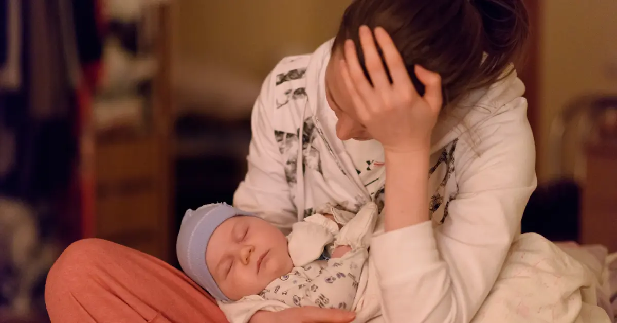 Mãe segura o bebê adormecido no colo enquanto apoia a cabeça com expressão cansada — uma imagem que revela a verdade por trás da culpa.