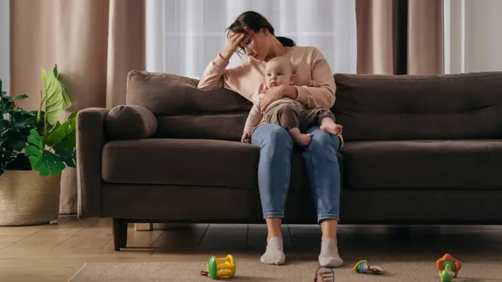Pessoa sentada em um sofá com expressão cansada, segurando uma criança no colo em meio a brinquedos espalhados — uma cena que revela a verdade por trás da maternidade.