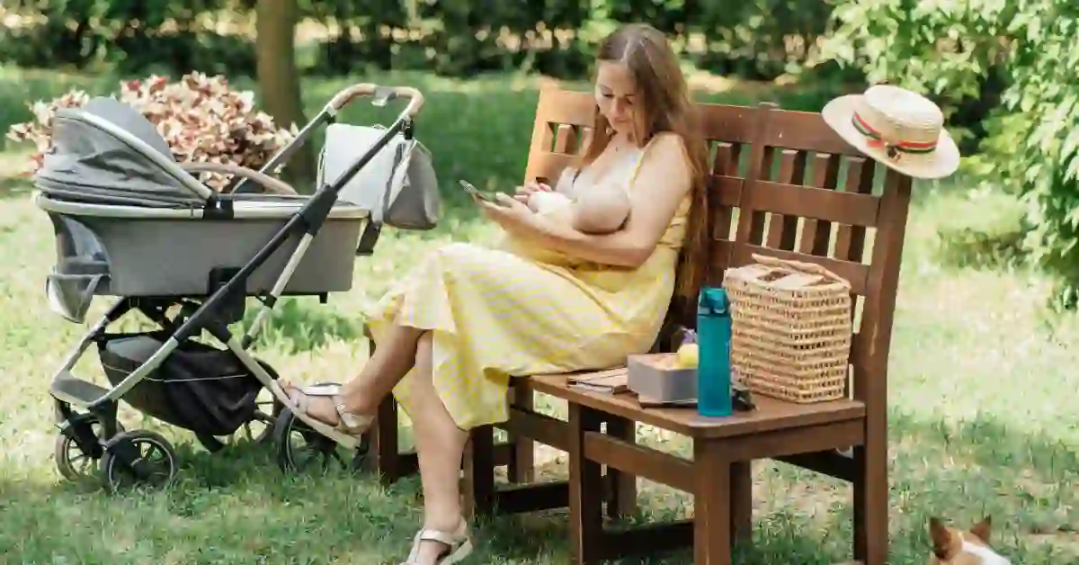 Mulher amamentando bebê em banco de parque enquanto usa o celular, possivelmente acessando um app de amamentação; ao lado, cesta de palha, garrafa de água e carrinho de bebê.