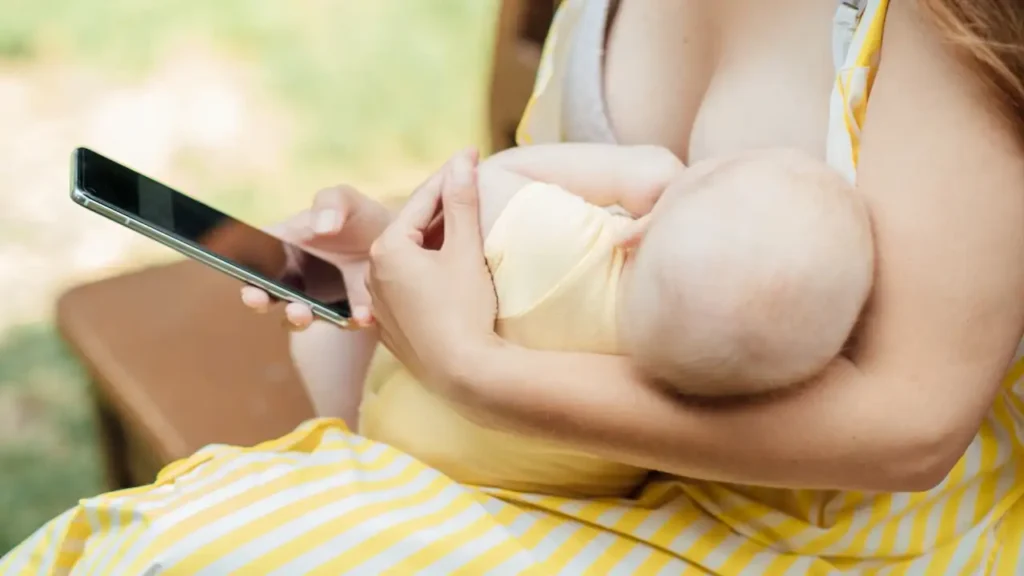 Mulher amamentando bebê vestido de amarelo enquanto usa o celular, possivelmente acessando um app de amamentação.