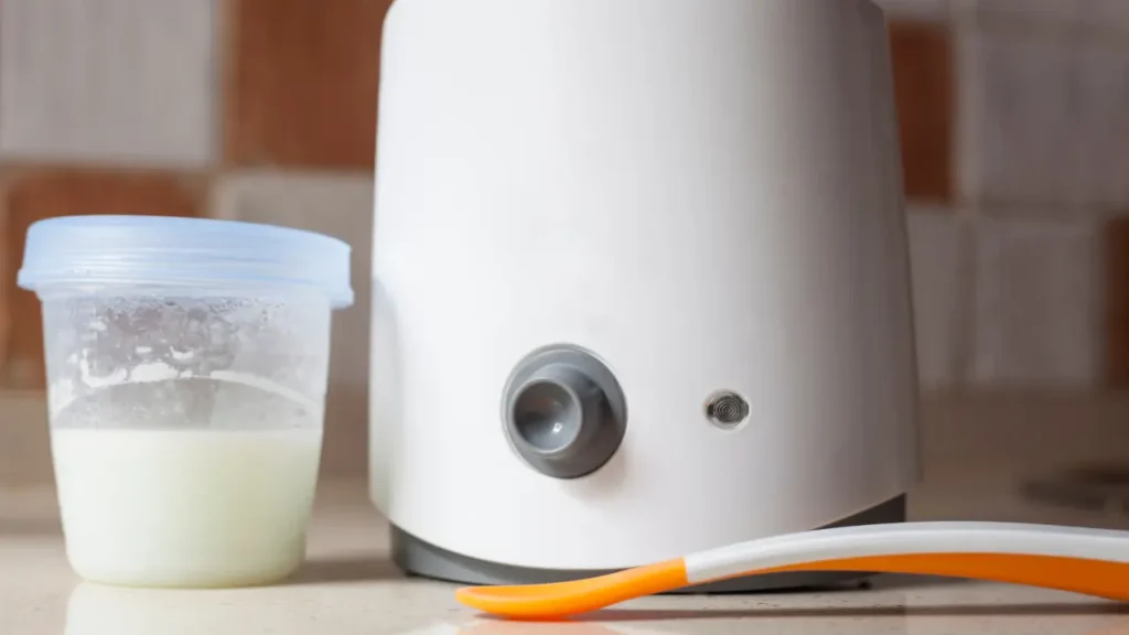 Aquecedor inteligente de mamadeira ao lado de recipiente com leite e colher infantil sobre bancada de cozinha.