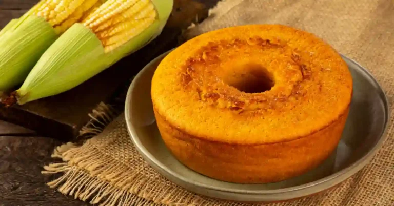 Bolo de milho de liquidificador redondo com furo central, servido em prato sobre tecido de juta, ao lado de espigas de milho frescas.