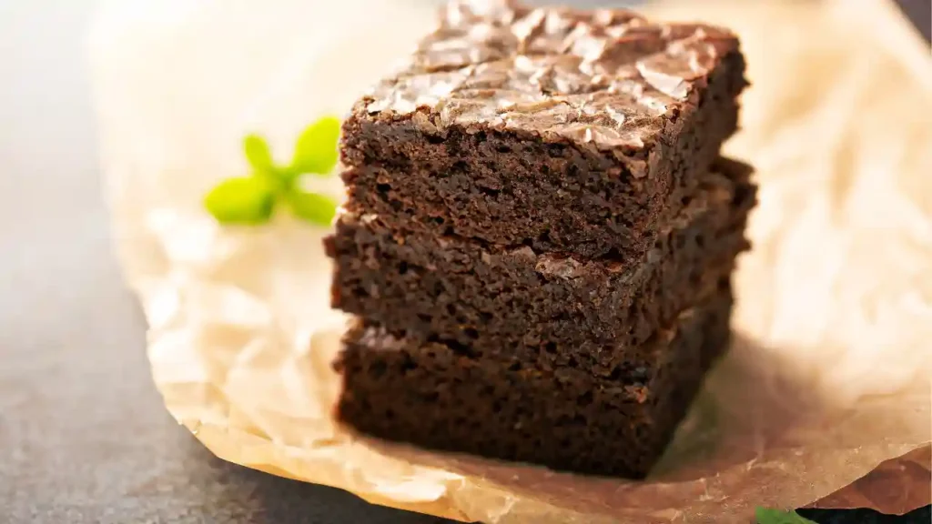 Três brownies fit e saudáveis empilhados sobre papel manteiga, com textura macia e folha de hortelã ao fundo.