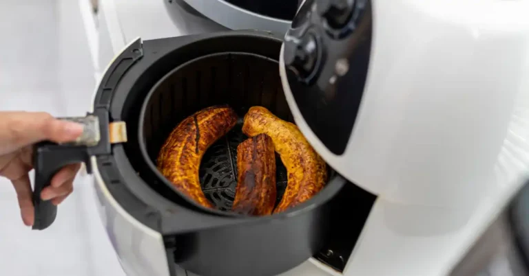Air fryer branca com três bananas-da-terra assadas, sendo puxada por uma mão — opção saudável de café da manhã fit na Air fryer.