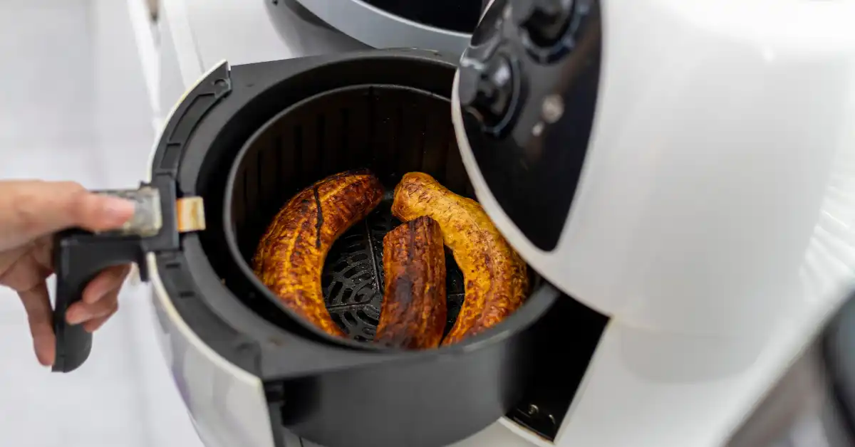 Air fryer branca com três bananas-da-terra assadas, sendo puxada por uma mão — opção saudável de café da manhã fit na Air fryer.