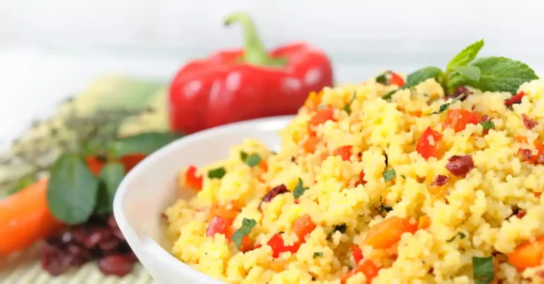 Cuscuz fit colorido servido em uma tigela branca, com legumes variados como pimentão vermelho, cebola roxa, abobrinha e uma azeitona preta no centro, decorado com ramo de salsa.