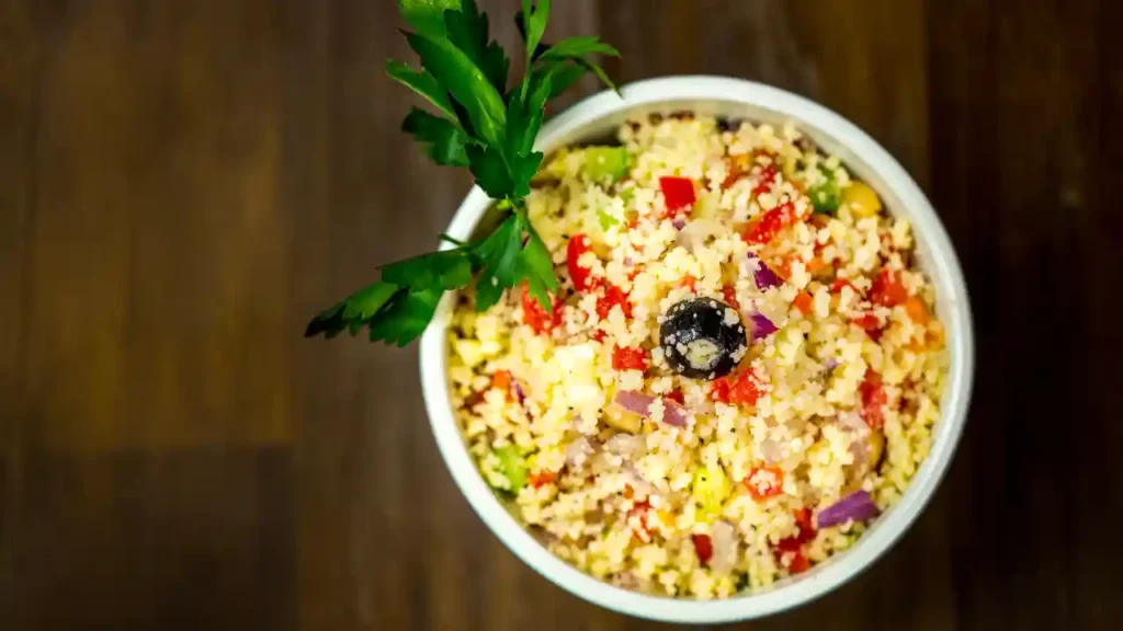 Cuscuz fit colorido servido em uma tigela branca, com legumes variados como pimentão vermelho, cebola roxa, abobrinha e uma azeitona preta no centro, decorado com ramo de salsa.