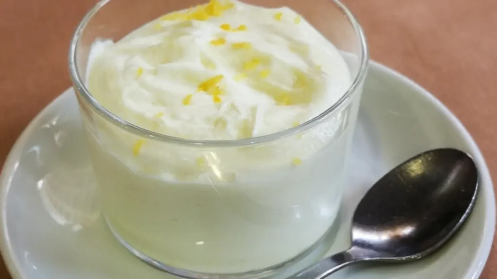 Doce gelado com leite em pó servido em um copo de vidro, decorado com raspas de limão, acompanhado de colher sobre pires branco.