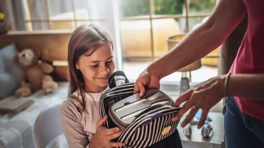 Criança sorridente segura uma lancheira listrada enquanto um adulto coloca um recipiente com talheres, possivelmente contendo pastel de banana, dentro da lancheira.