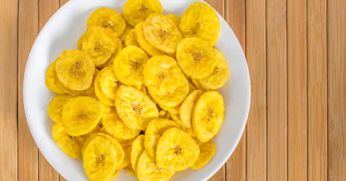 Prato branco com várias rodelas crocantes de pastel de banana sobre uma superfície de madeira clara.