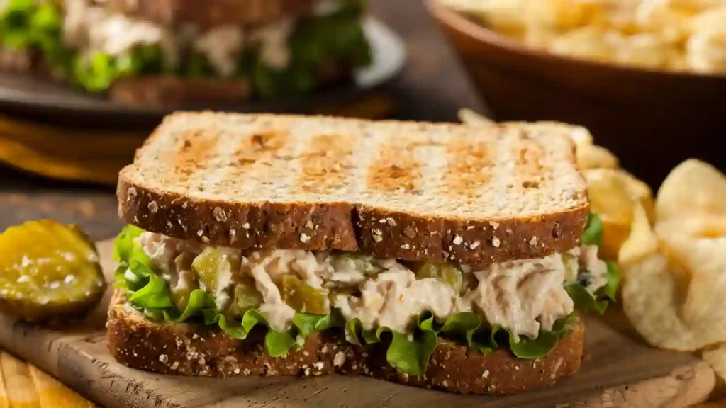 Sanduíche de salada de atum com picles e alface em pão integral, servido sobre uma tábua de madeira com chips e fatia de picles ao fundo, ideal para Lanches rápidos para o dia a dia.