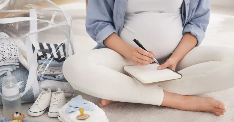 Pessoa grávida sentada no chão escrevendo em um caderno, ao lado de itens de bebê como roupas, mamadeira e sapatinhos — momento de organização com foco na lista do que levar na maternidade.