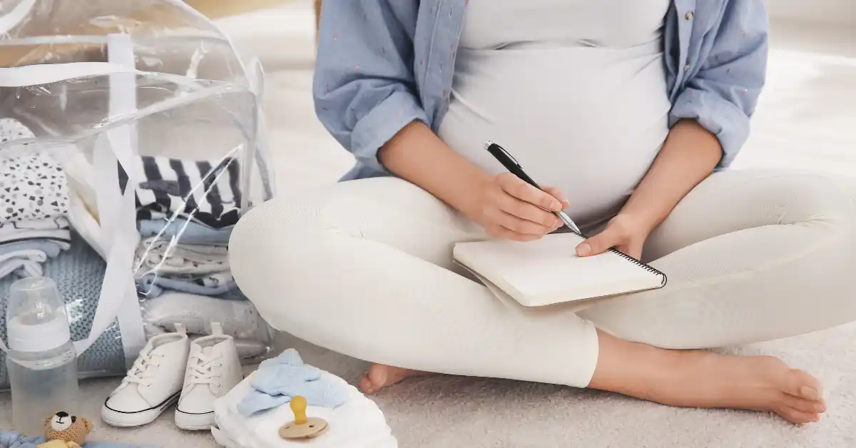 Pessoa grávida sentada no chão escrevendo em um caderno, ao lado de itens de bebê como roupas, mamadeira e sapatinhos — momento de organização com foco na lista do que levar na maternidade.